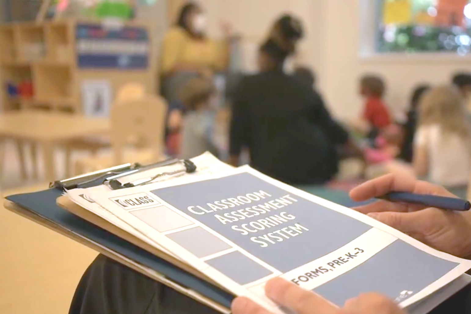 A male observer sits in the foreground with the class score sheet in focus. In the background, you can see the blurred images of a teacher sitting on the carpet with children. This is a still photo pulled from the New E3 School in Norfolk VA.