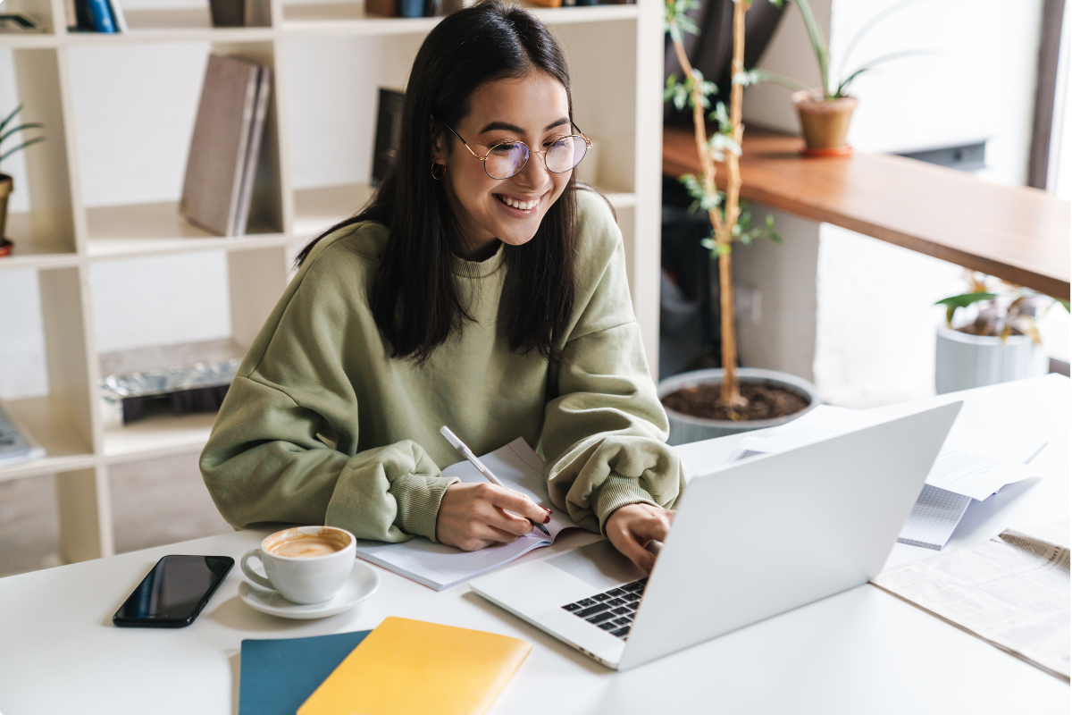 Teacher with glasses sitting at table in front of laptop, smiling, typing, writing in notebook