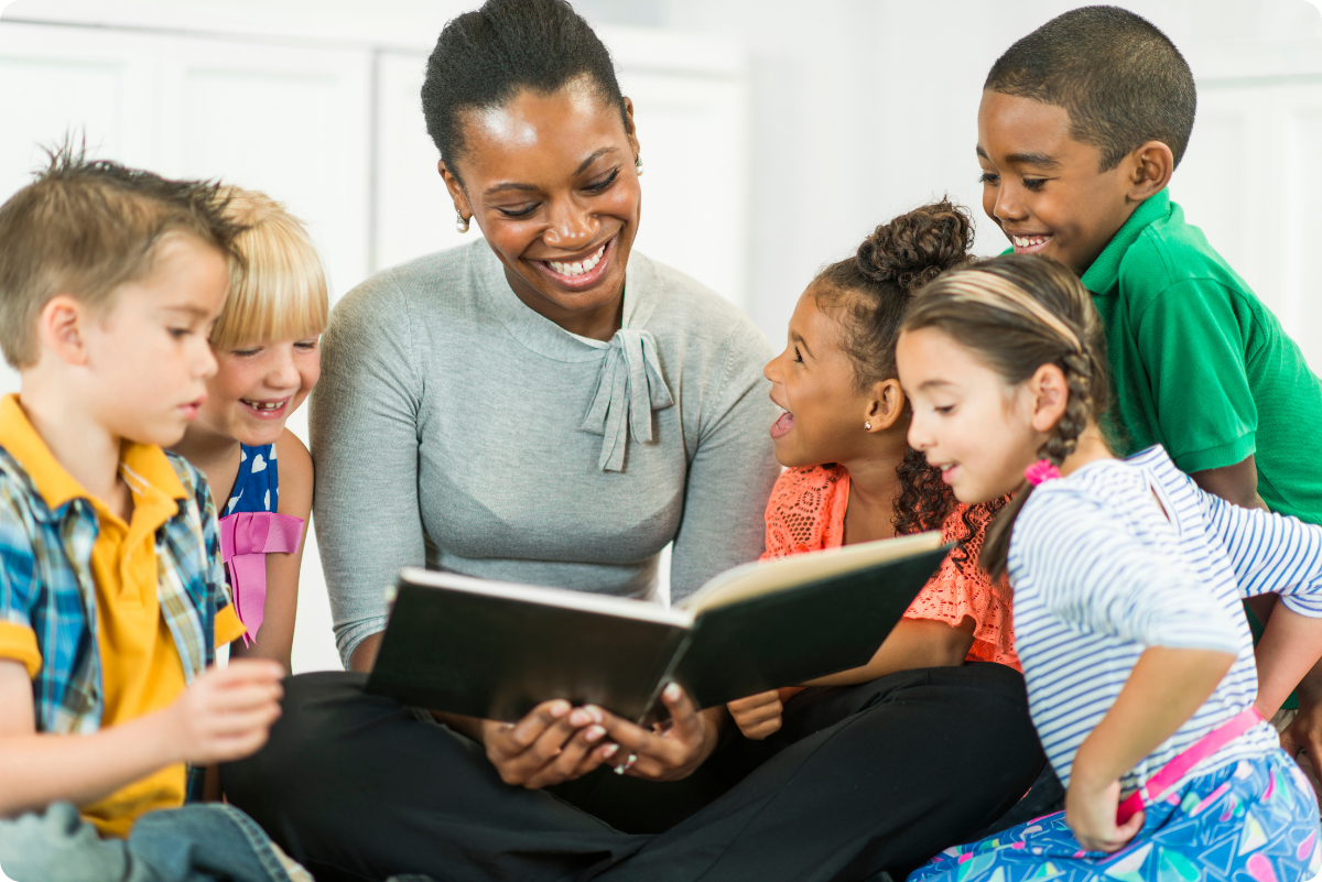 A preschool educator is sitting and smiling with children on the classroom floor as she reads to them.